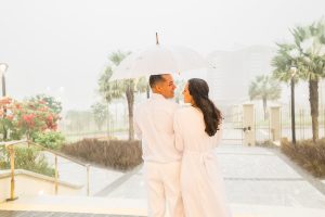 Noivos de costas sob muita chuva no templo de Fortaleza. estao segurando um guarda chuva branco.