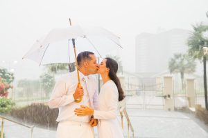 Noivos se beijando, no templo de Fortaleza, sob muita chuva. Noivo segurando guarda chuva branco