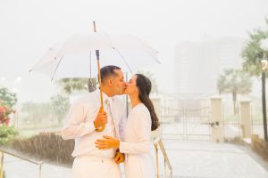 Noivos se beijando, no templo de Fortaleza, sob muita chuva. Noivo segurando guarda chuva branco