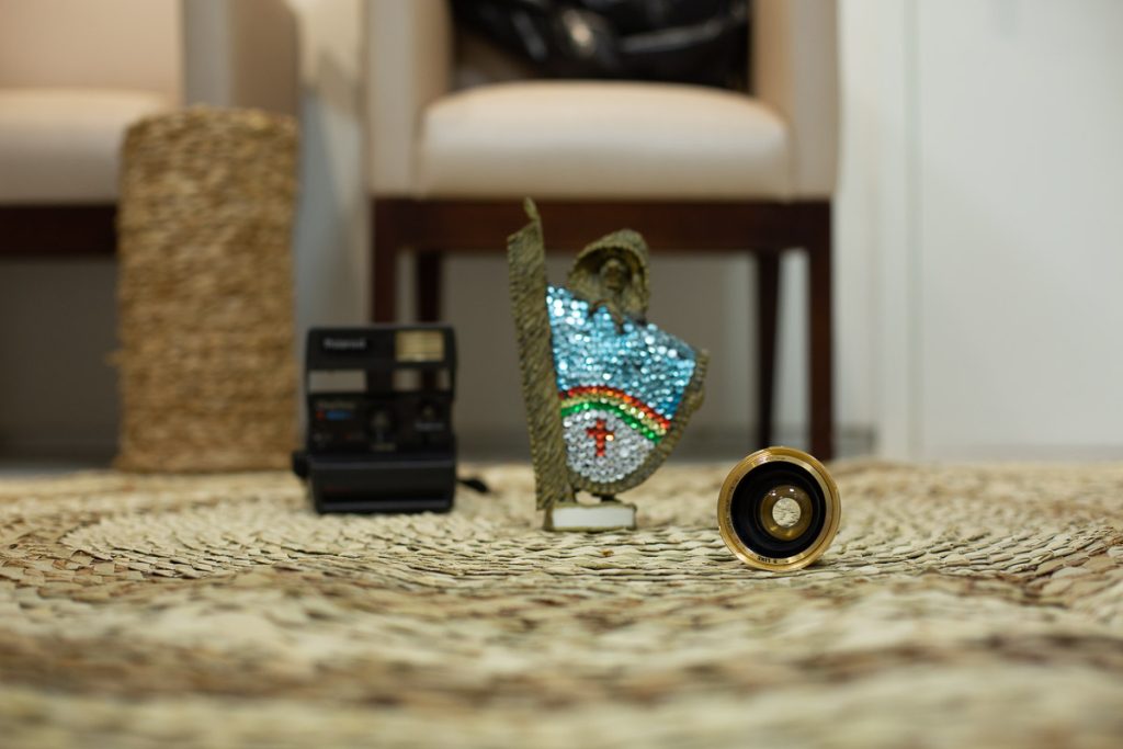 foco numa lente de maquina de fotografia antiga, sobre tapete de palha com fundo desfocado, onde da para perceber uma cadeira, um vaso, uma polaroide antiga e um caboclo rural de enfeite com as cores do estado de pernambuco.