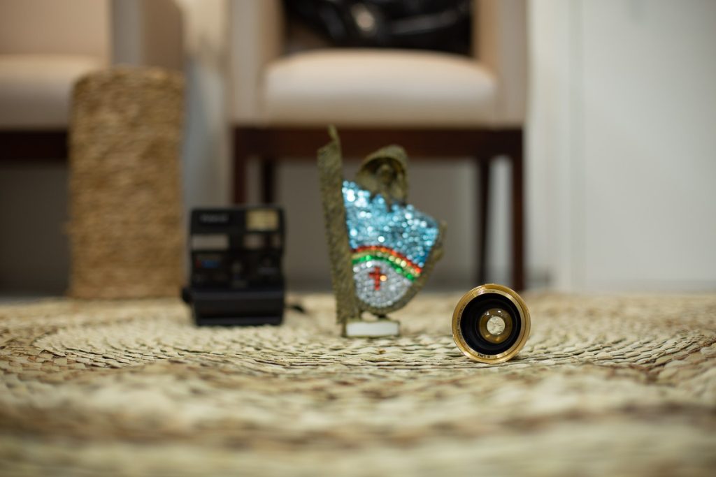 foco numa lente de maquina de fotografia antiga, sobre tapete de palha com fundo desfocado, onde da para perceber uma cadeira, um vaso, uma polaroide antiga e um caboclo rural de enfeite com as cores do estado de pernambuco.