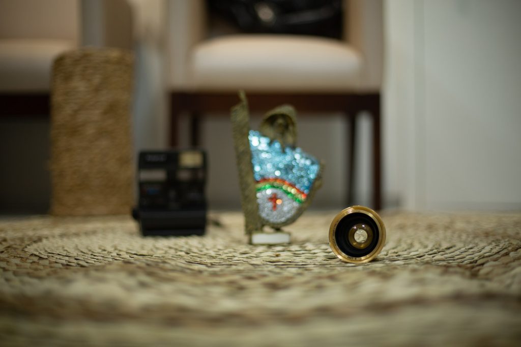 foco numa lente de maquina de fotografia antiga, sobre tapete de palha com fundo desfocado, onde da para perceber uma cadeira, um vaso, uma polaroide antiga e um caboclo rural de enfeite com as cores do estado de pernambuco.