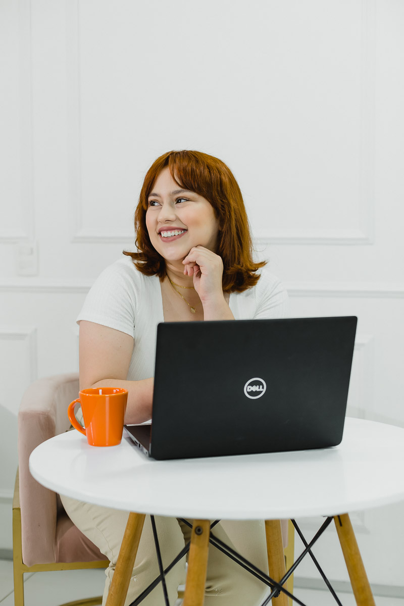 executiva branca de cabelos ruivos trabalhando ao celular e computador. está com roupas claras e uma xícara laranja, cadeira rosa e fundo branco