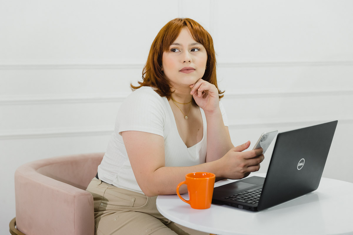 executiva branca de cabelos ruivos trabalhando ao celular e computador. está com roupas claras e uma xícara laranja.