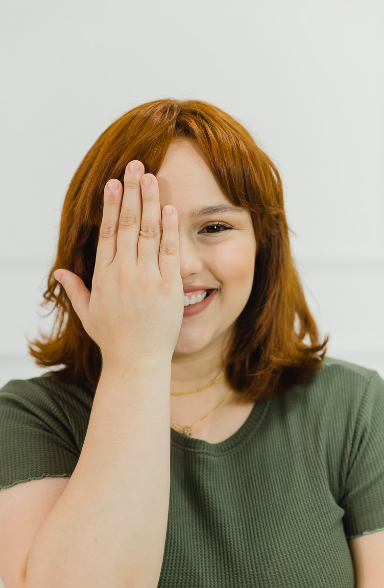 mulher branca de cabelos ruivos, com camiseta verde tapando um dos olhos com a mão e sorrindo.