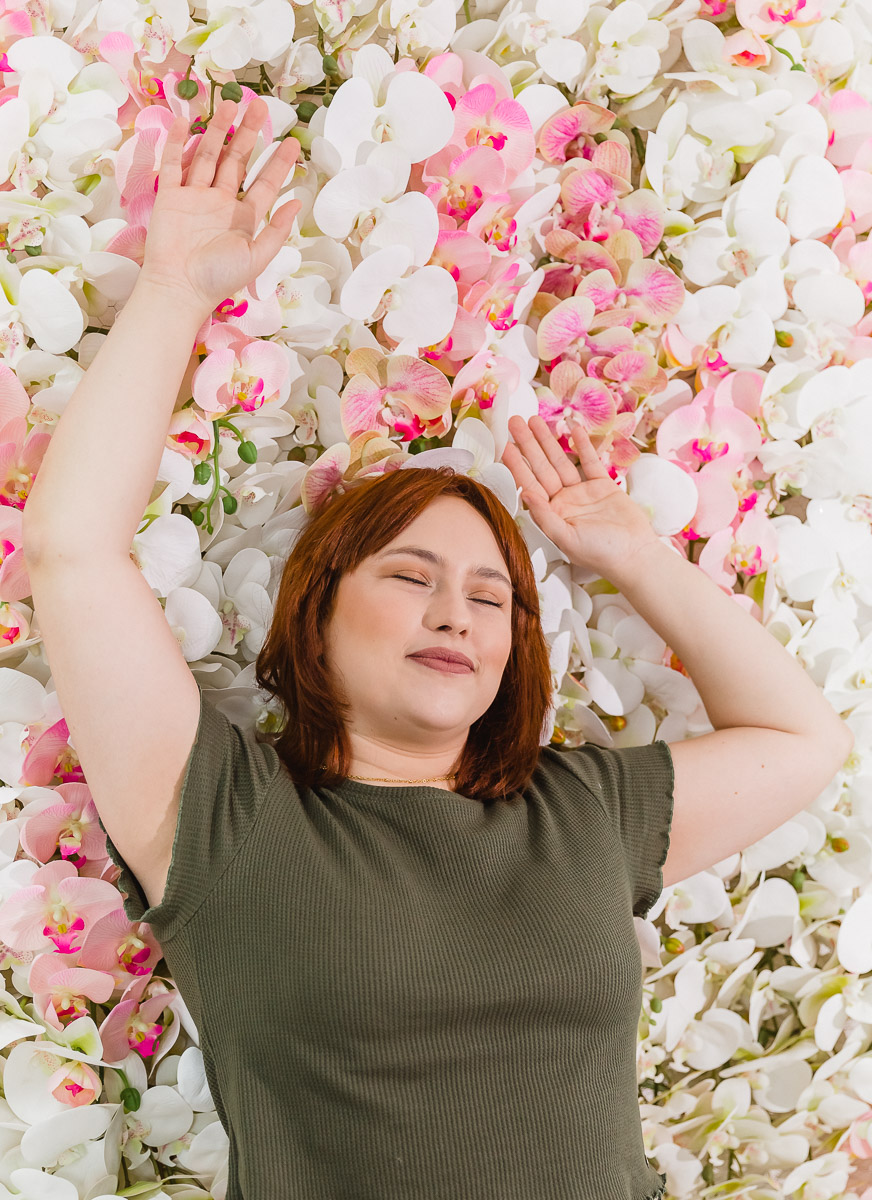 mulher branca de cabelos ruivos deitada com olhos fechados e sorridente  sobre fundo altamente florido