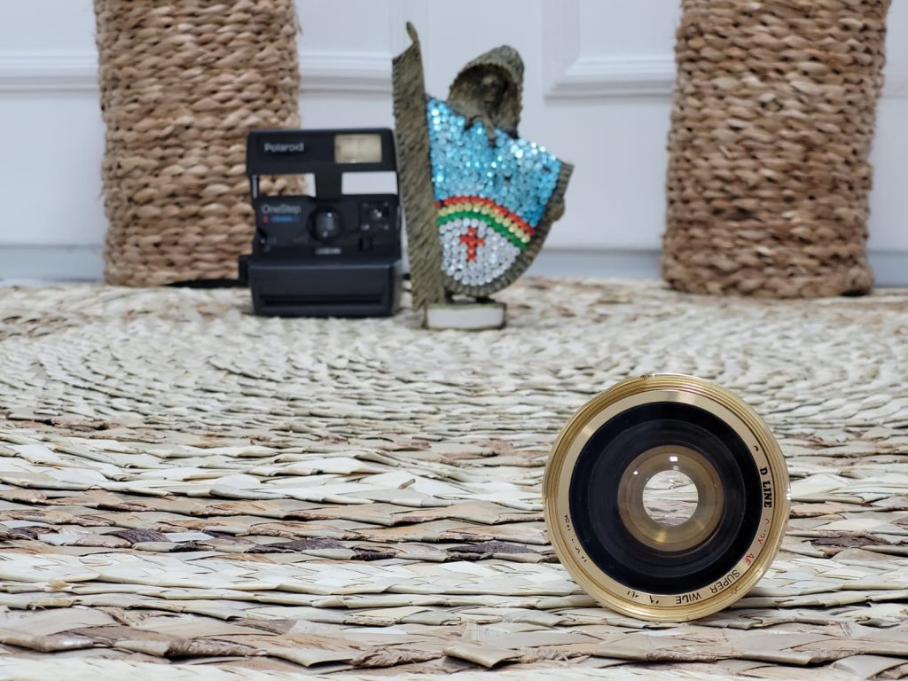 lente de maquina de fotografia antiga bem no primeiro plano, sobre tapete de palha com fundo desfocado, onde da para perceber, um vaso, uma polaroide antiga e um caboclo rural de enfeite com as cores do estado de pernambuco.