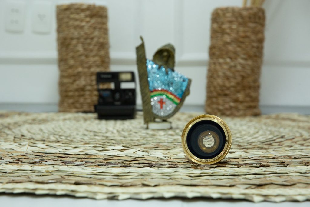 lente de maquina de fotografia antiga bem no primeiro plano, sobre tapete de palha com fundo desfocado, onde da para perceber, um vaso, uma polaroide antiga e um caboclo rural de enfeite com as cores do estado de pernambuco.