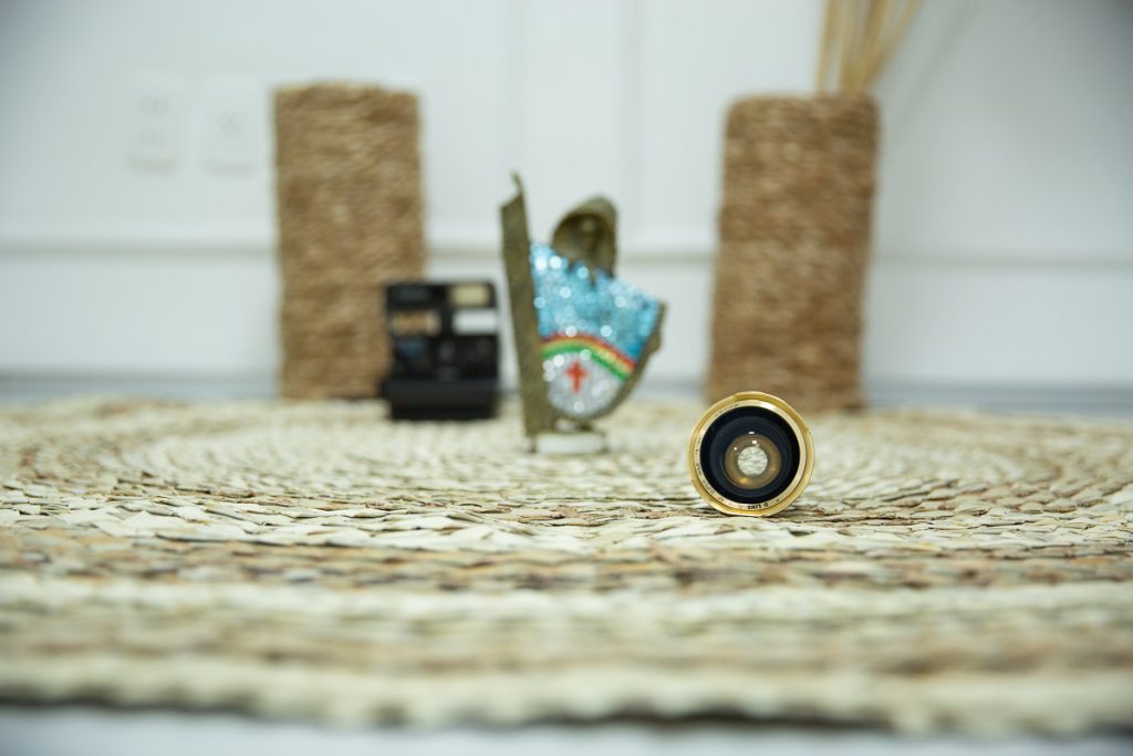 foco numa lente de maquina de fotografia antiga, sobre tapete de palha com fundo desfocado, onde da para perceber uma cadeira, um vaso, uma polaroide antiga e um caboclo rural de enfeite com as cores do estado de pernambuco.