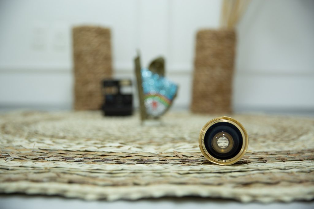 foco numa lente de maquina de fotografia antiga, sobre tapete de palha com fundo desfocado, onde da para perceber, um vaso, uma polaroide antiga e um caboclo rural de enfeite com as cores do estado de pernambuco.