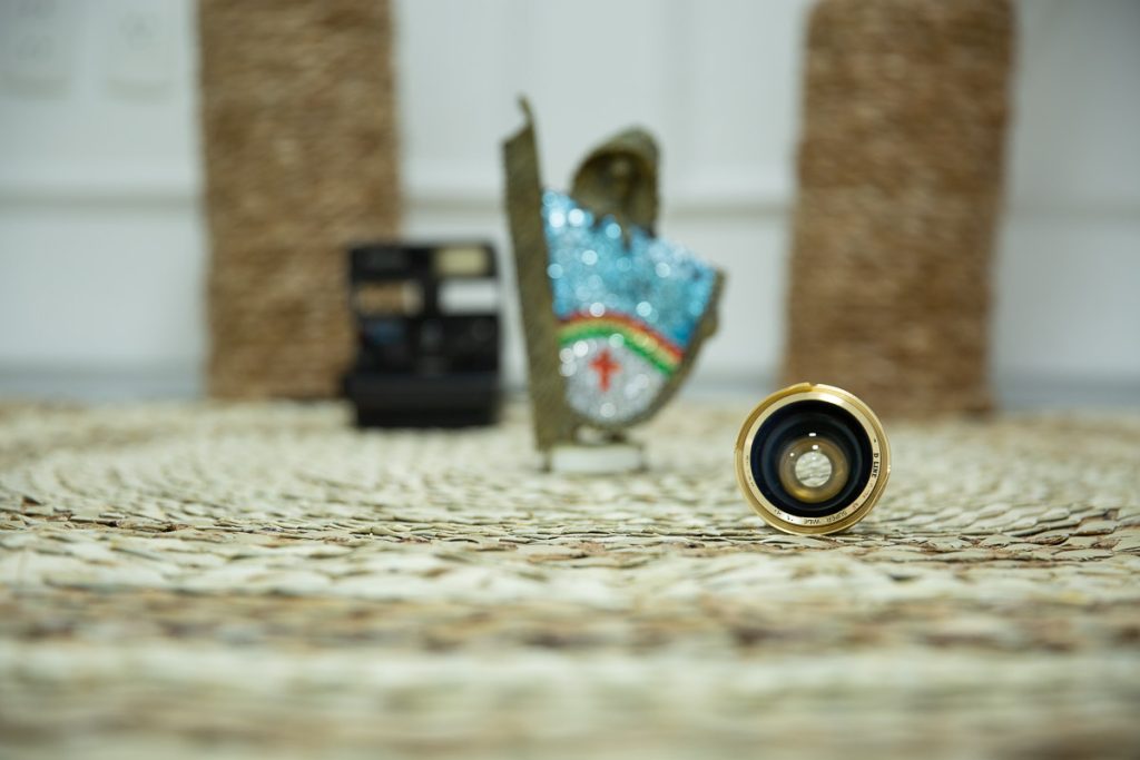 lente de maquina de fotografia antiga bem no primeiro plano, sobre tapete de palha com fundo desfocado, onde da para perceber, um vaso, uma polaroide antiga e um caboclo rural de enfeite com as cores do estado de pernambuco.