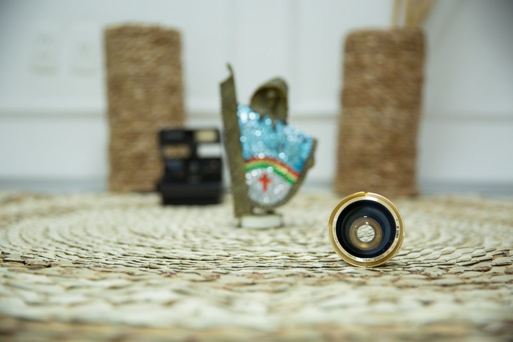 lente de maquina de fotografia antiga bem no primeiro plano, sobre tapete de palha com fundo desfocado, onde da para perceber, um vaso, uma polaroide antiga e um caboclo rural de enfeite com as cores do estado de pernambuco.