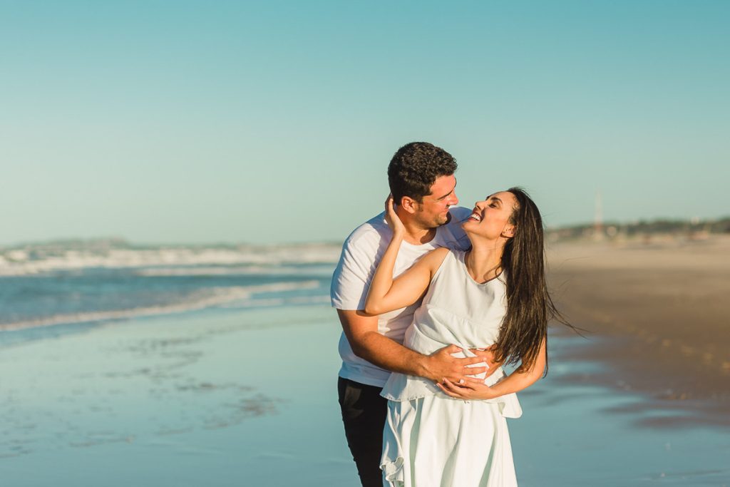 Casal sorridente e felizes, vestidos de branco se abraçando em pé, na beira da praia com o céu azul ao fundo.