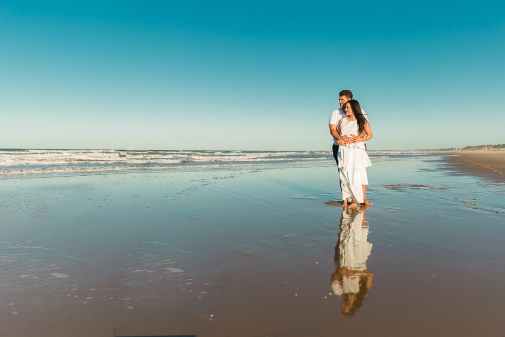 Casal vestidos de branco se abraçando, sorridentes em pé, na beira da praia com o céu azul ao fundo e o reflexo no espelho de agua