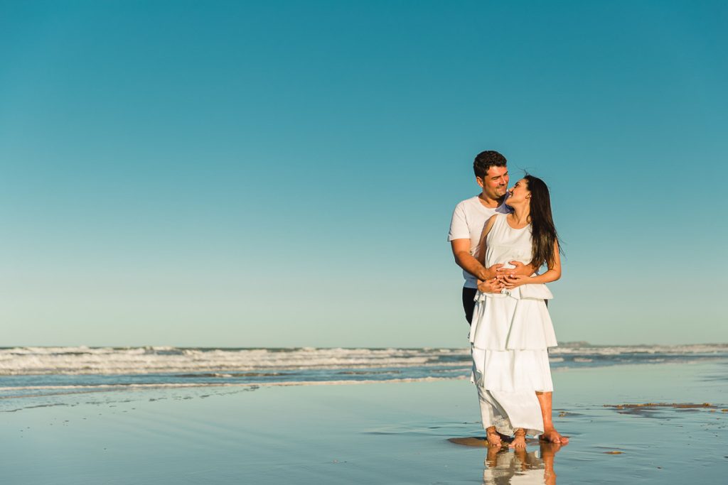 Casal sorridente e felizes, vestidos de branco se abraçando entre olhares em pé, na beira da praia com o céu azul ao fundo.