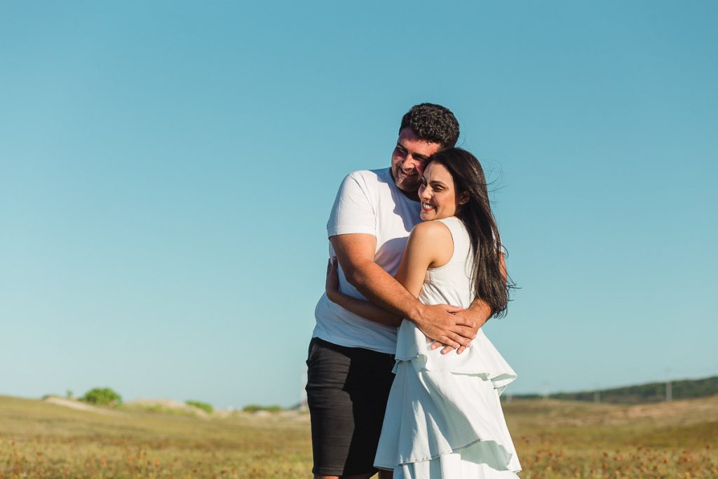 Casal sorridente e felizes, vestidos de branco se abraçando em pé no gramado rústico com o céu azul ao fundo.