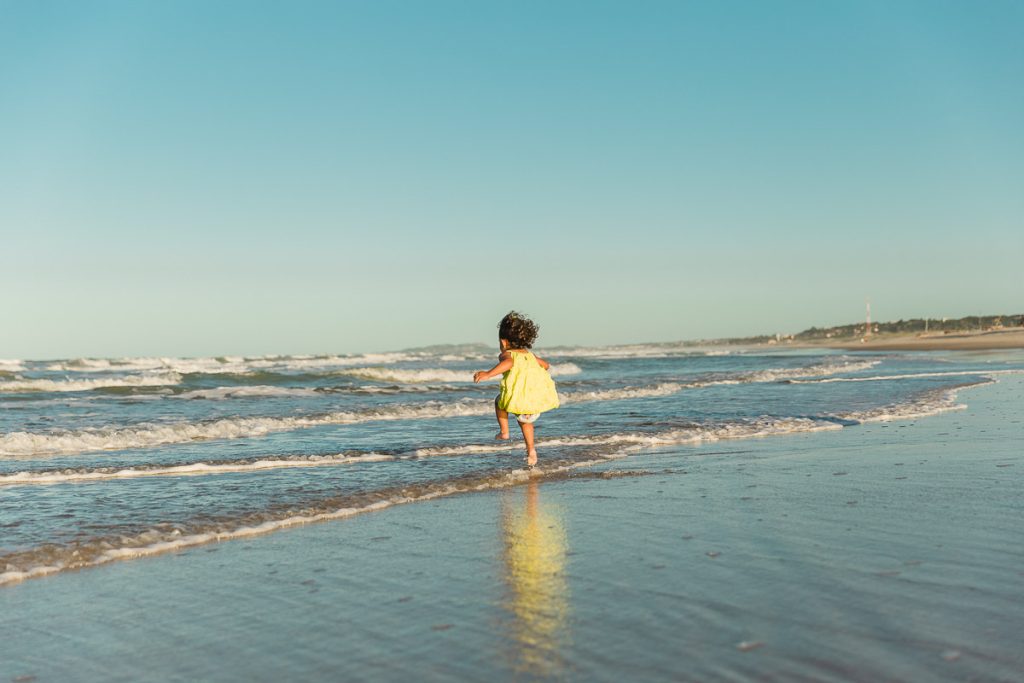 Menina de 6 anos, vista de costas pulando nas ondas da praia. Céu azul ao fundo.
