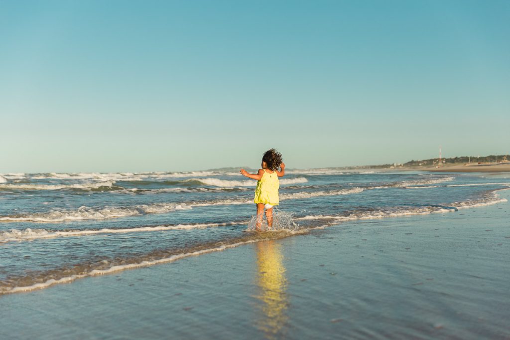 Menina de 6 anos, vista de costas, caindo em pé nas ondas da praia. Céu azul ao fundo.
