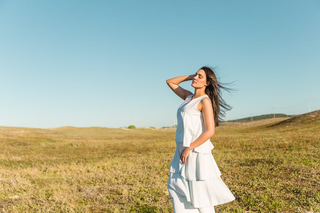 Mulher magra e bonita, vestida em branco com os cabelos voando, em pé no gramado rústico com o céu azul ao fundo.